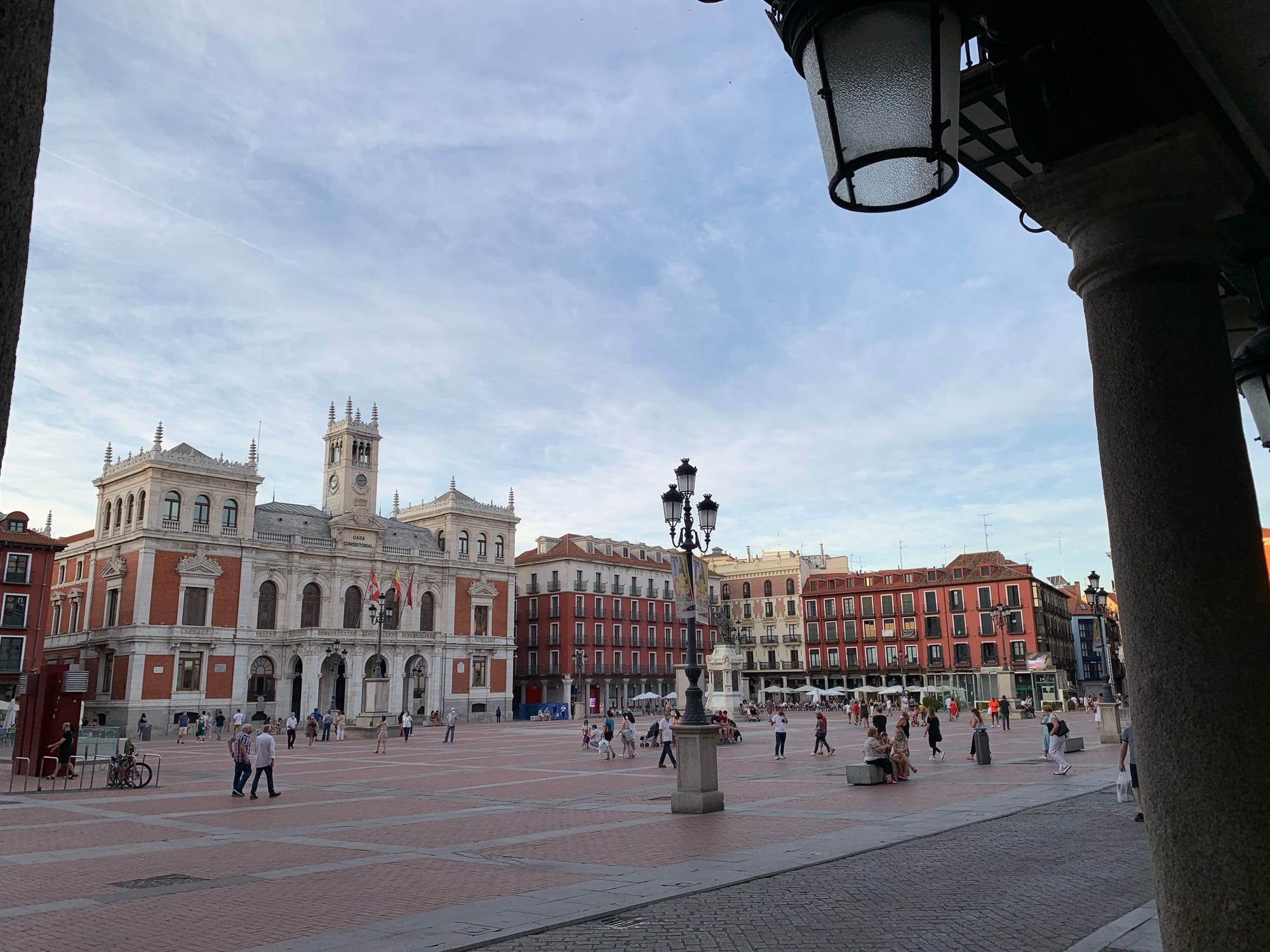 The Plaza Mayor of Valladolid seen from under its arcades on a summer evening: the Casa Consistorial, a white-stone and red-brick town hall with a central clock tower and turreted corner pavilions, stands on the left, rows of red-painted arcaded houses close the far side, an ornate iron lamp post rises from the wide red-and-grey paved square where people stroll, and a granite column and hanging lantern of the arcade frame the right edge under a pale blue sky streaked with cloud