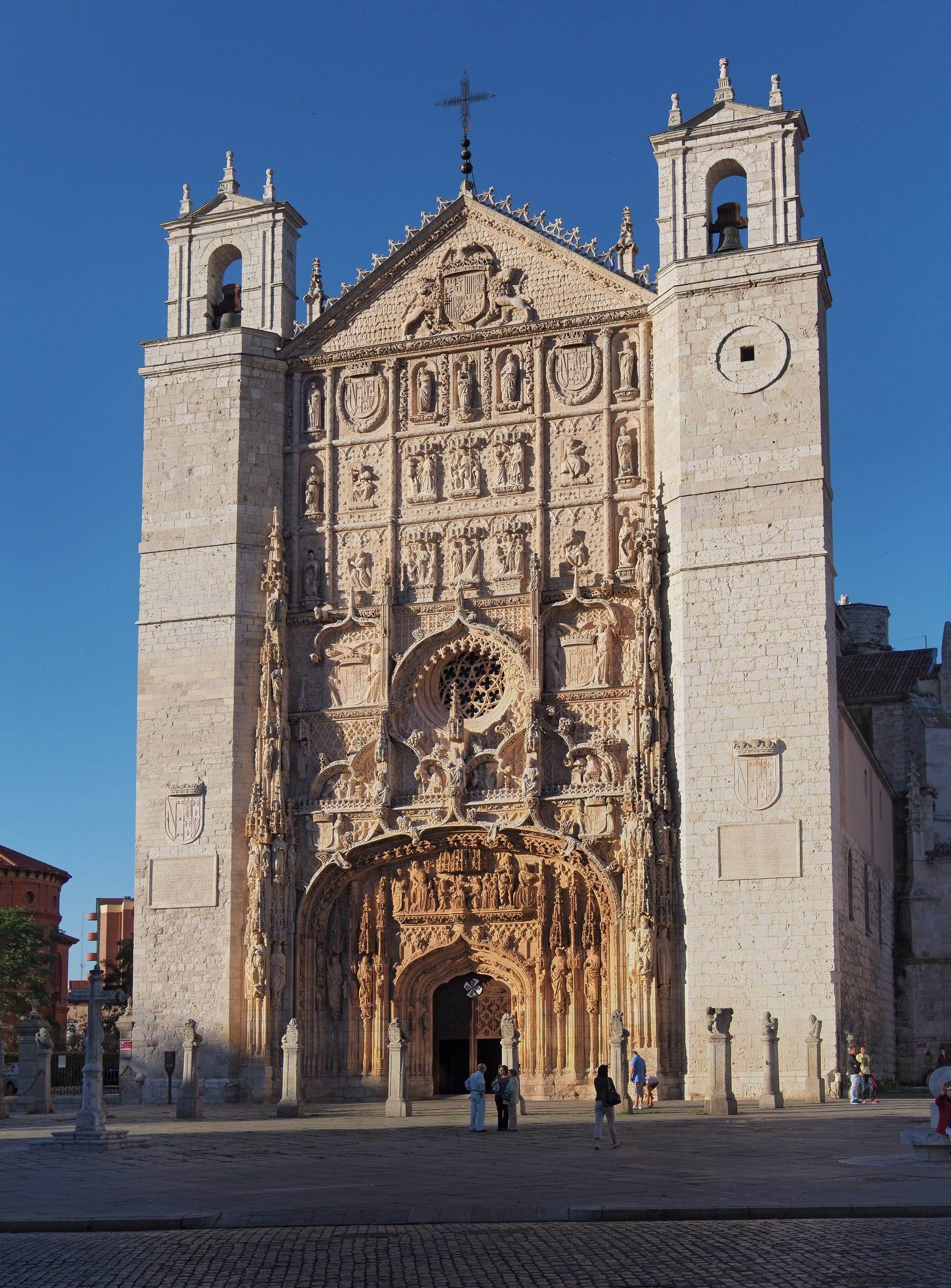 The façade of the church of San Pablo in Valladolid in warm evening light: between two plain stone towers topped by belfries, the Isabelline Gothic frontispiece is carved from the pointed gable down to the deep arched portal with tiers of saints in niches, royal coats of arms, pinnacles, tracery and a rose window, the sculpted portal glowing golden, with a stone wayside cross and a row of stone posts on the square in front under a deep blue sky