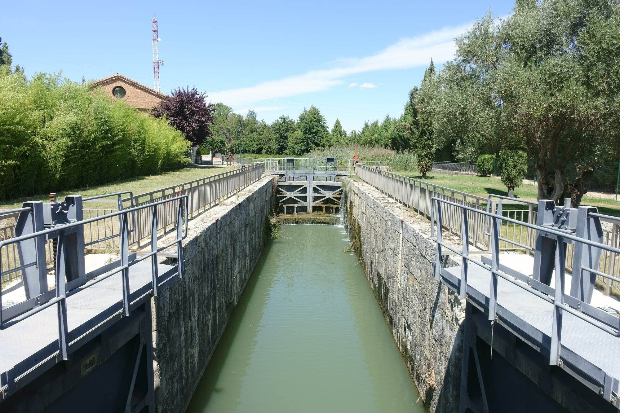 Lock 42 of the Canal de Castilla in Valladolid, the last lock before the canal basin, seen from its lower gates: a deep, narrow chamber of grey dressed stone holds green water, closed at the far end by tall grey gates braced with diagonal beams, a thin stream spilling down from the higher level behind them, while steel footbridges with railings and gate winches cross the near end between lawns, an olive tree and a red-brick building under a blue summer sky