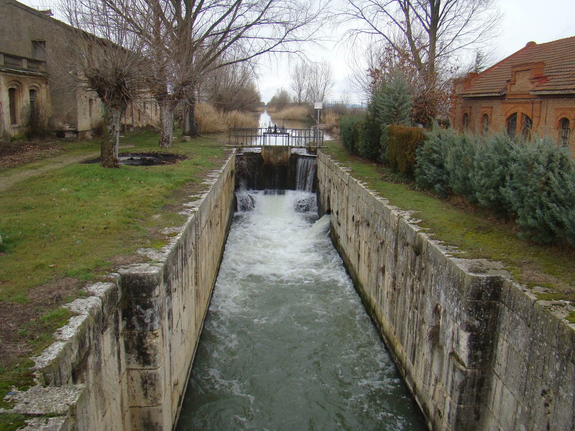 Lock 40 of the Canal de Castilla at Corcos, in the province of Valladolid, on a grey winter day: water from the upper reach pours over and through the closed gate at the far end into a deep chamber of weathered limestone blocks and churns white as it runs toward the viewer, while beyond the gate's iron footbridge the canal continues straight between reeds and bare trees, flanked by an old stone building and a red-brick house